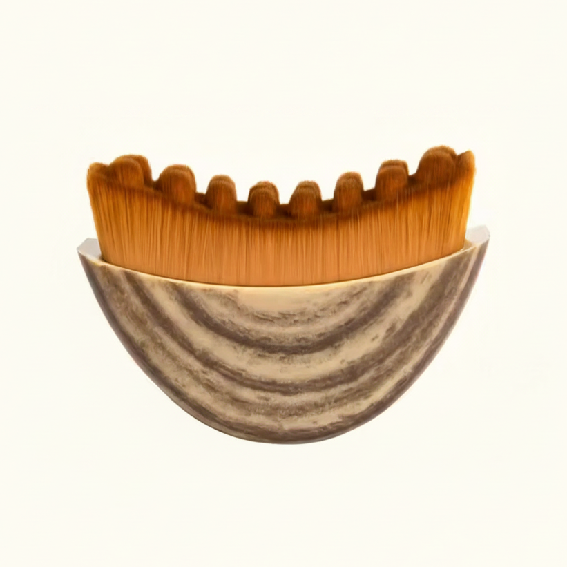 A close-up of a makeup brush with dense orange-brown bristles and a marbled beige and gray bowl-like base, shown against a plain white background.
