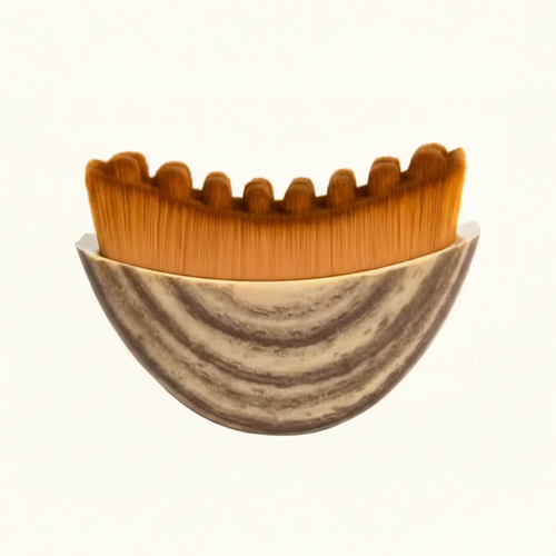 A close-up of a makeup brush with dense orange-brown bristles and a marbled beige and gray bowl-like base, shown against a plain white background.